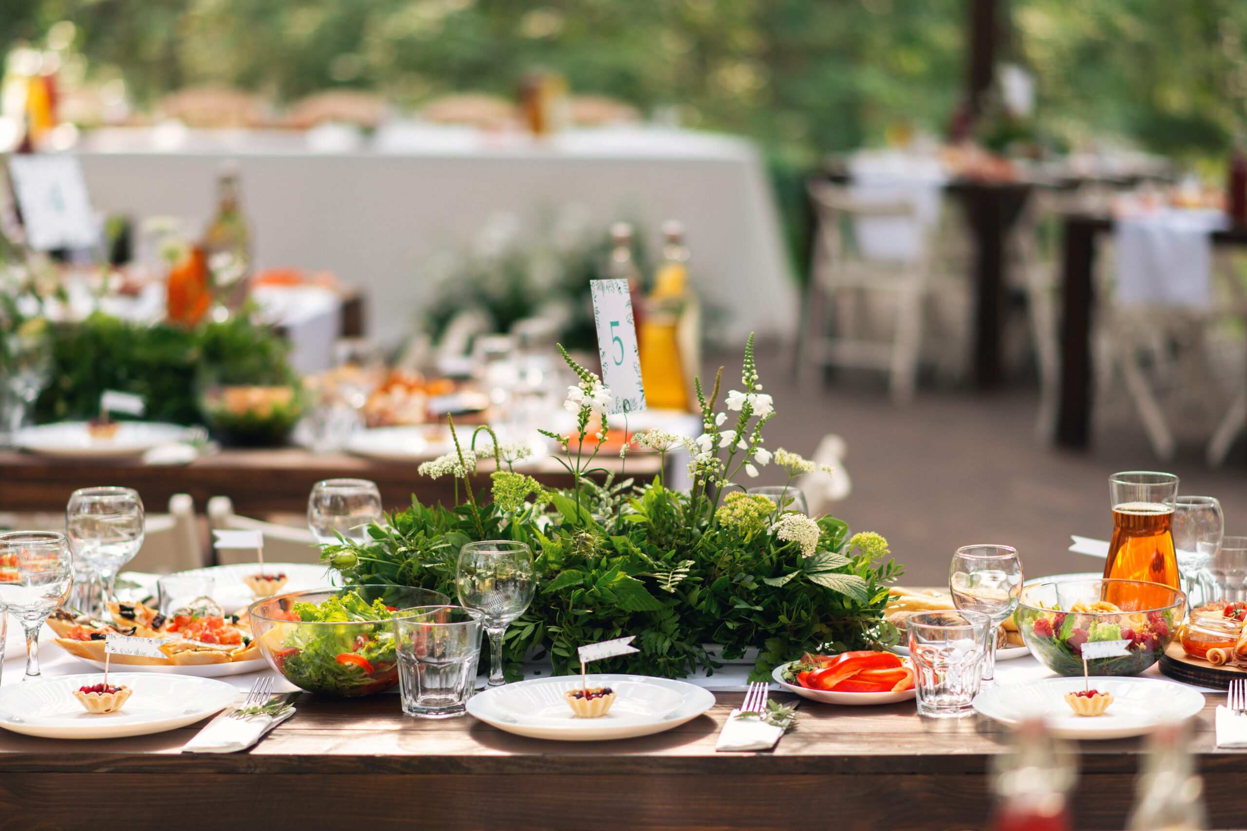 Outdoor banquet table set with salads, appetizers, floral centerpieces, and table number five at a garden event.