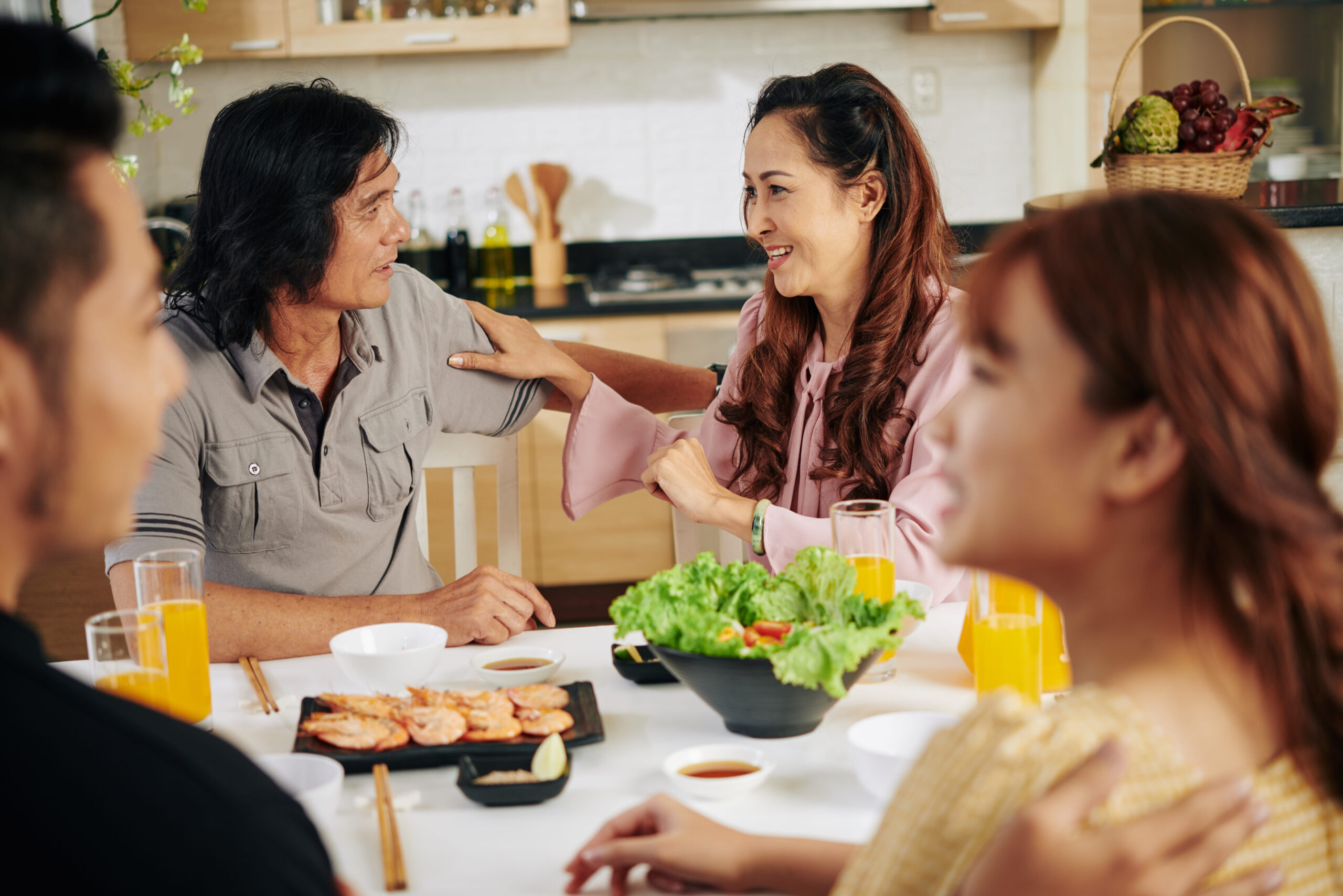 Older Asian couple smiling and talking with family during a healthy dinner in a bright, modern kitchen.