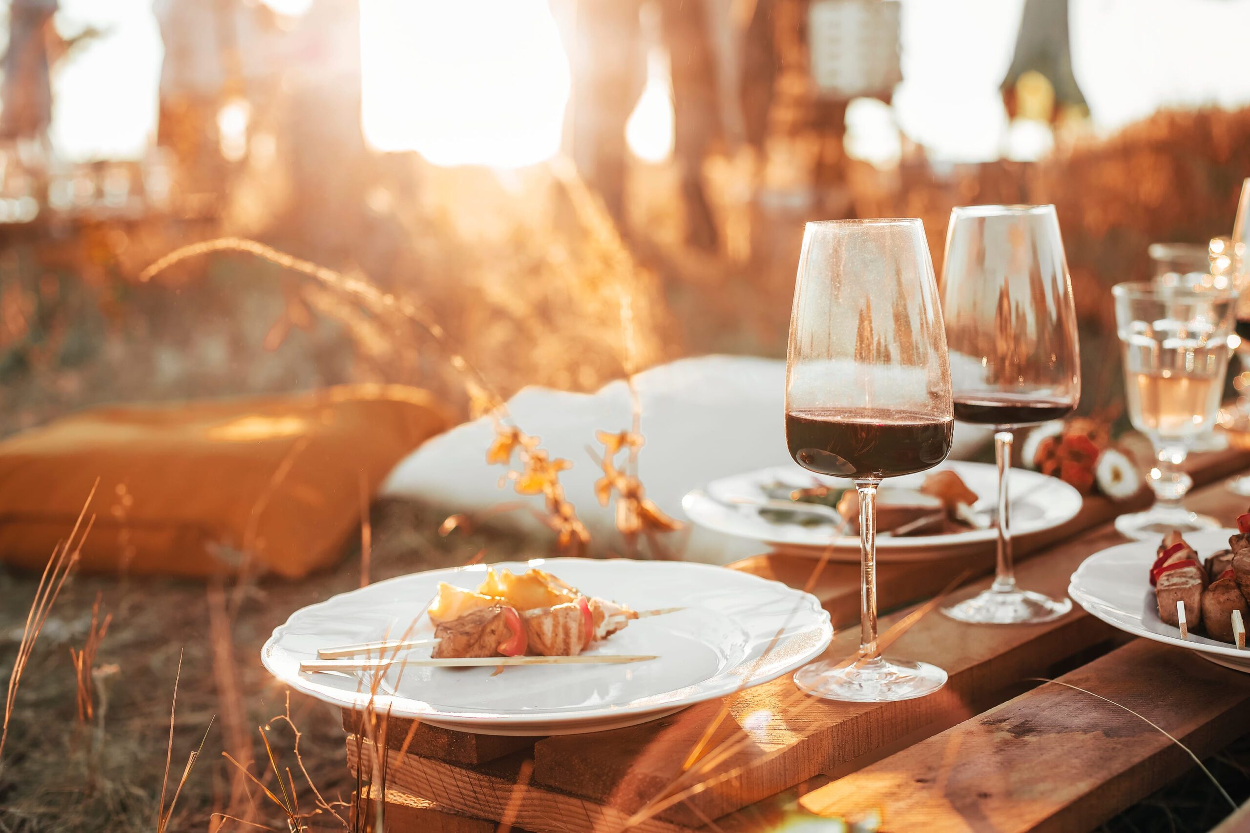 Two glasses of red wine and grilled vegetable skewers on a wooden table at a sunset outdoor picnic.