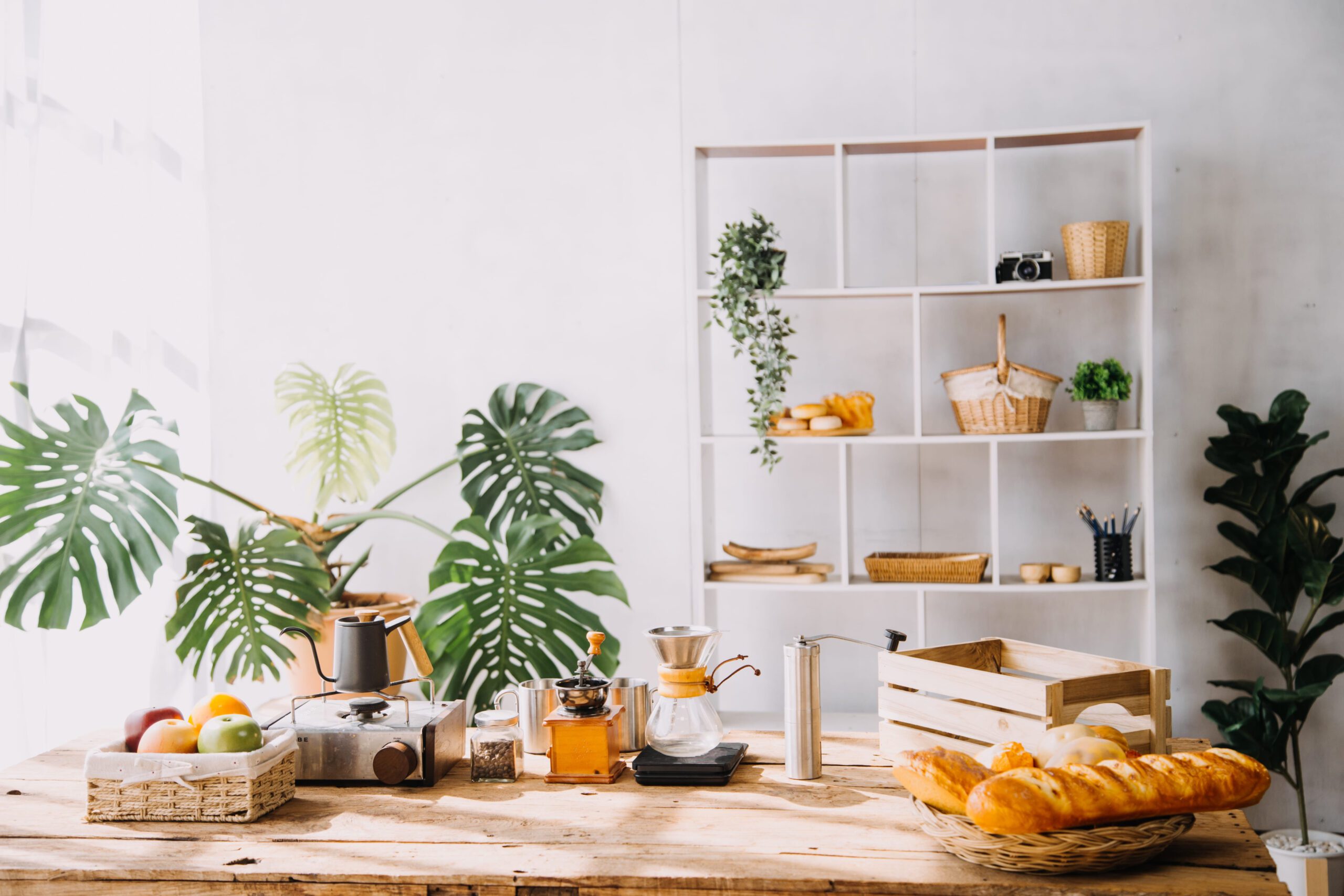 Manual coffee making equipment, fresh baguettes, and fruit on a wooden table in a bright, plant-filled room.