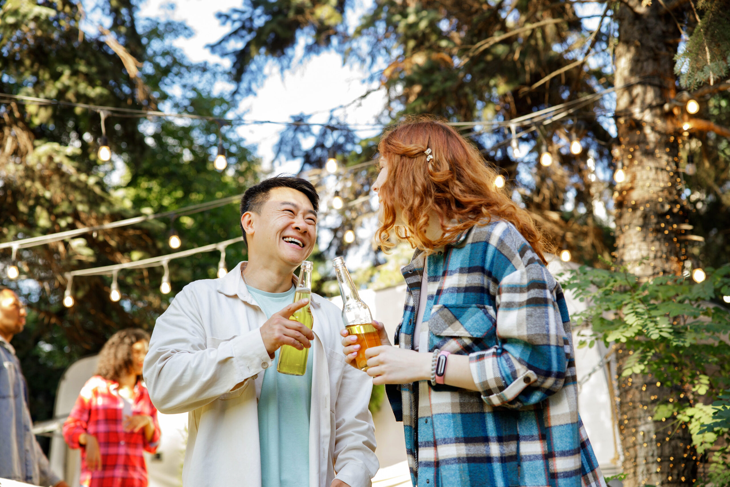 A man and a red-haired woman laughing and holding bottled drinks at an outdoor gathering under string lights.