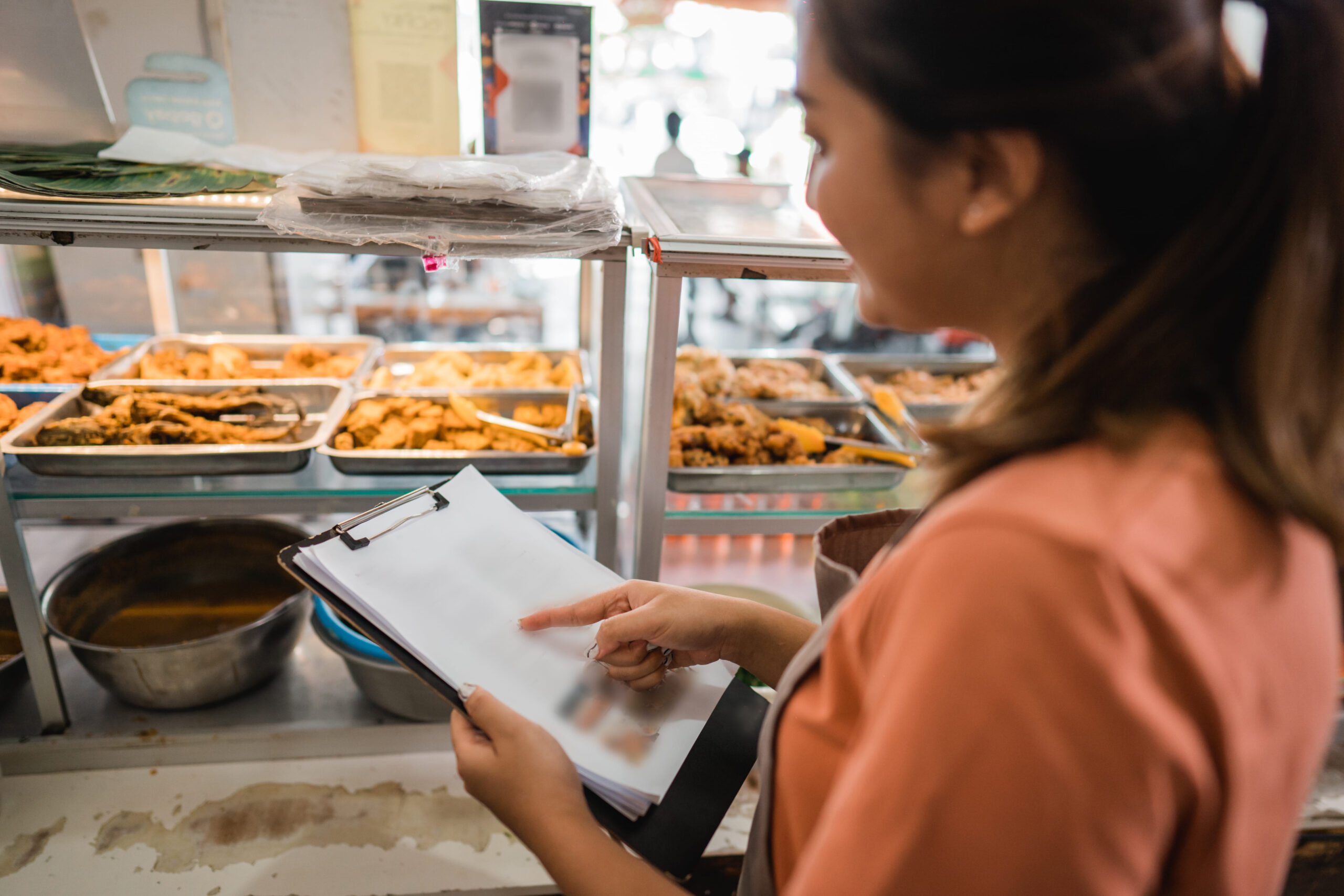 Female food stall owner checking a clipboard inventory in front of a glass display case filled with cooked dishes.