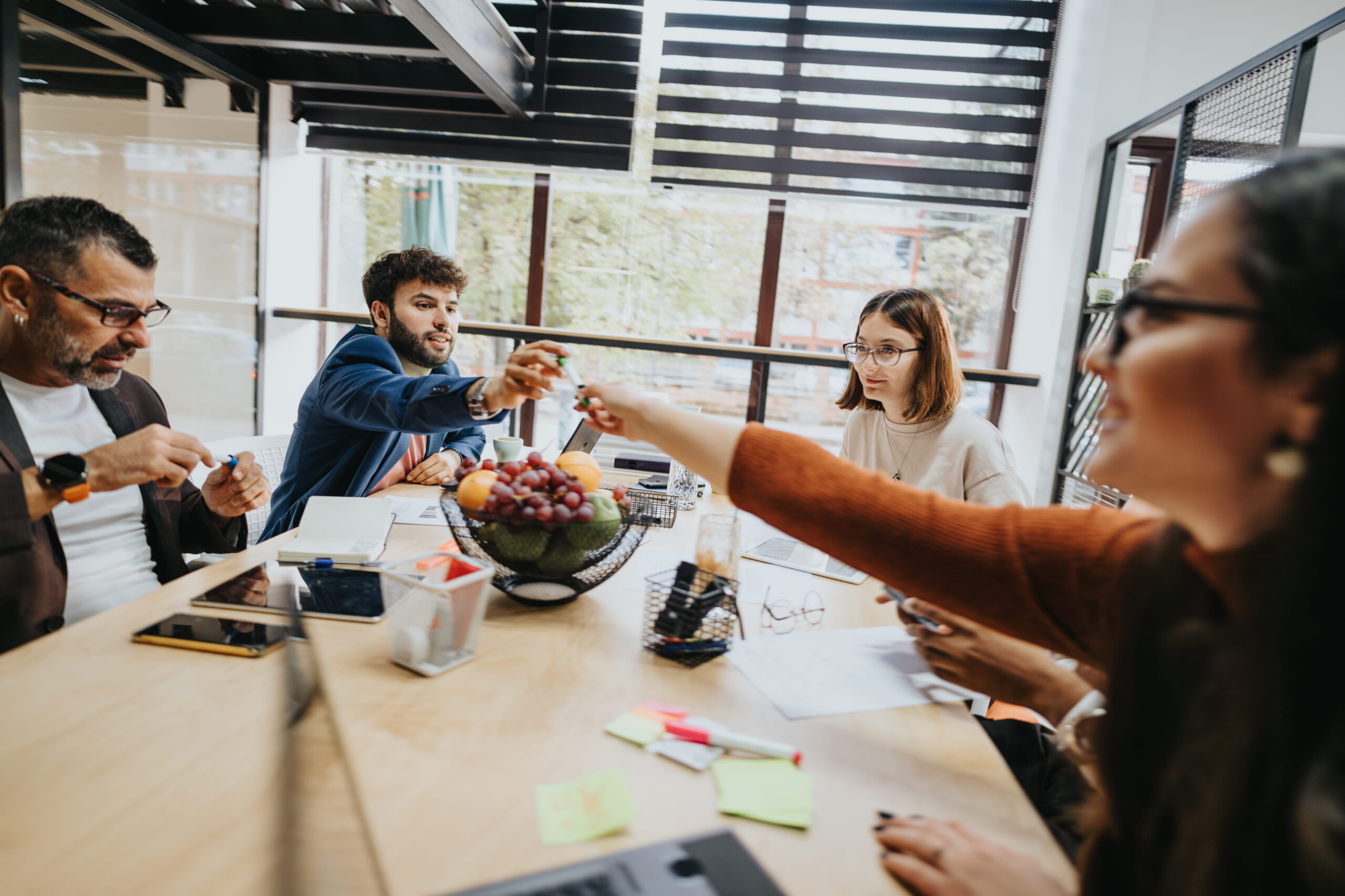 A diverse team of colleagues collaborating around a wooden table in a bright, modern office with fruit and stationery.