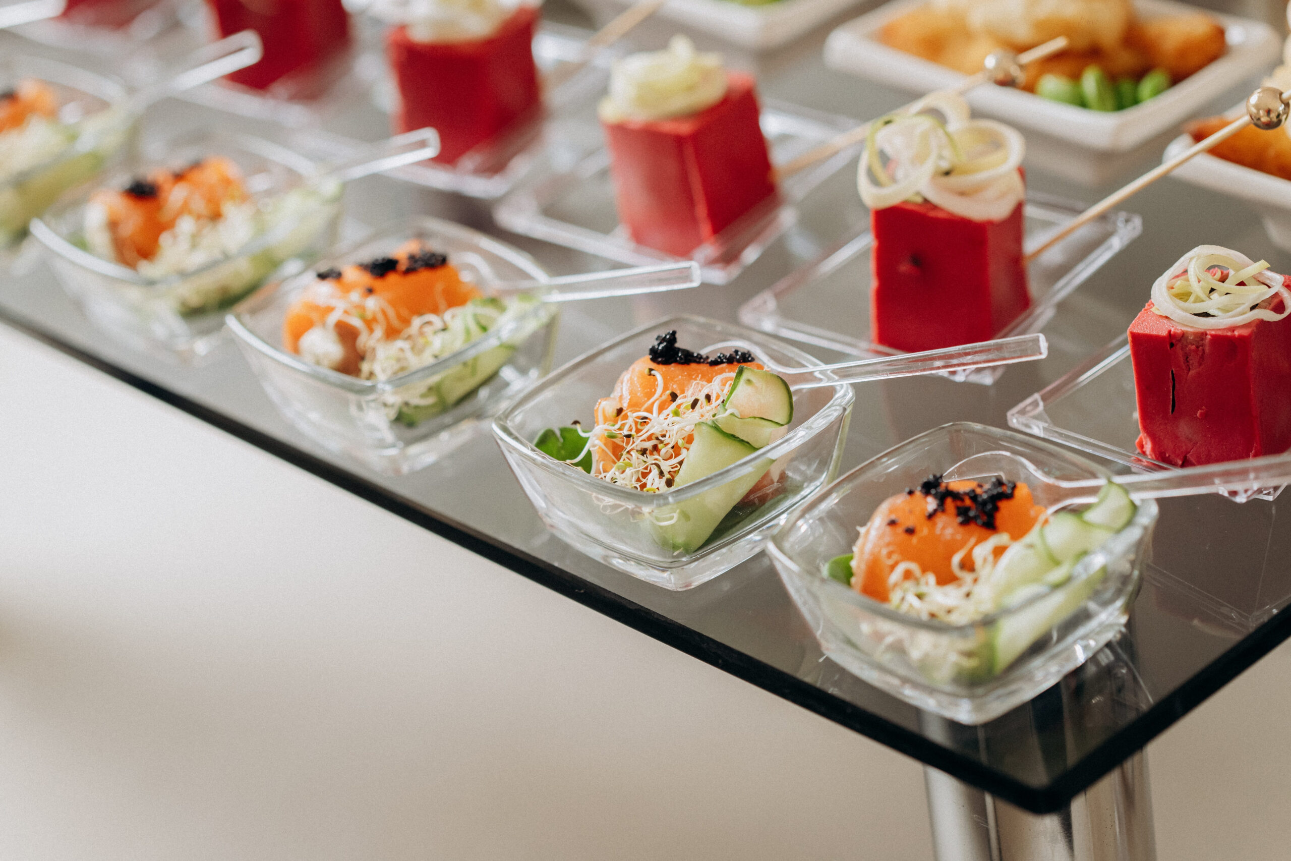 Gourmet catering appetizers with salmon and sprouts in glass bowls next to beet-colored mousse squares on a glass table.