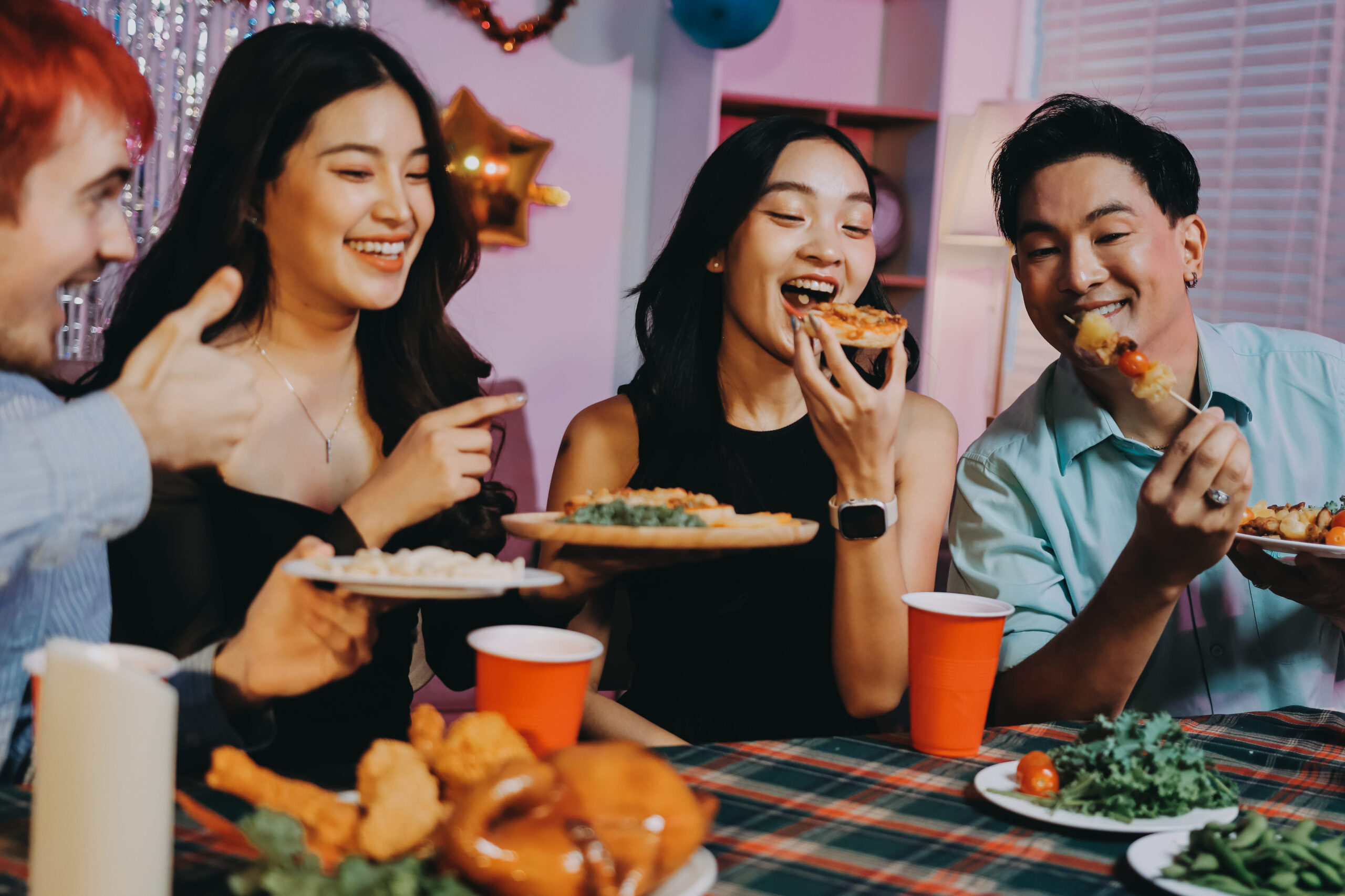 Four young friends laughing and eating pizza and skewers around a table at a festive dinner party.