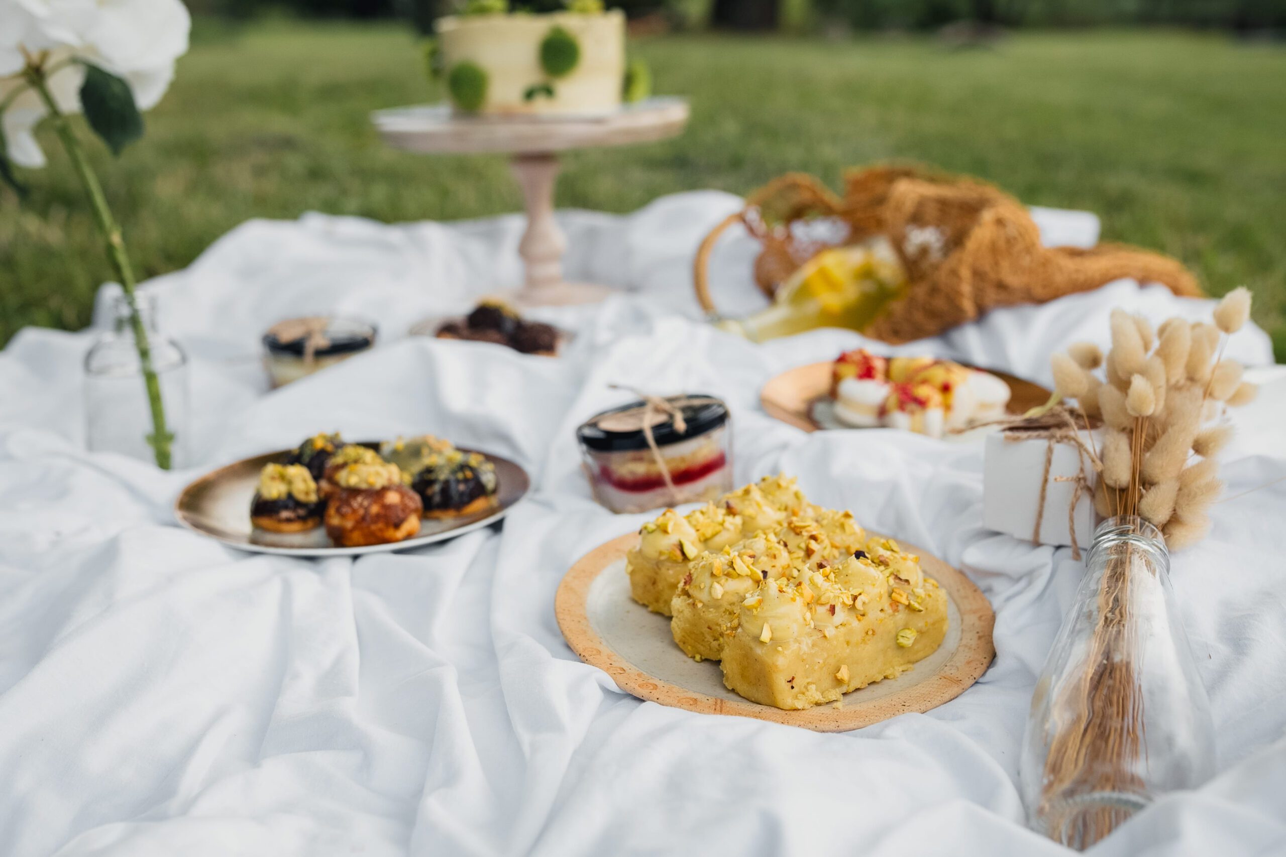 Assorted gourmet desserts including pistachio cake and pastries laid out on a white blanket for an outdoor picnic.